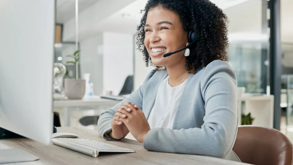 Woman sat at a desk with a computer. She has a headset on smiling, while giving customer service.